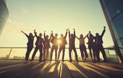 Silhouettes Of A Diverse Group Of Businesspeople Celebrating Success With Arms Raised On A Rooftop.